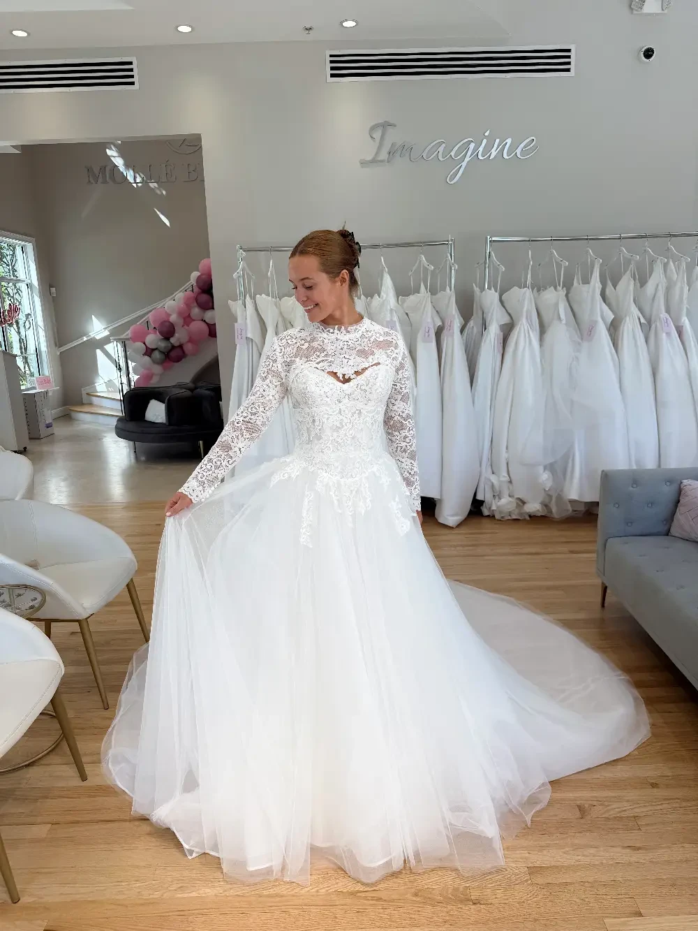 A woman in a wedding dress with long sleeves and lace details, standing in a bridal shop filled with white dresses.