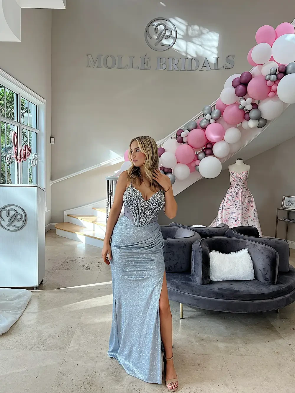 A woman wearing a silver gown with a slit stands in a bridal shop decorated with balloons and a wedding dress in the background.