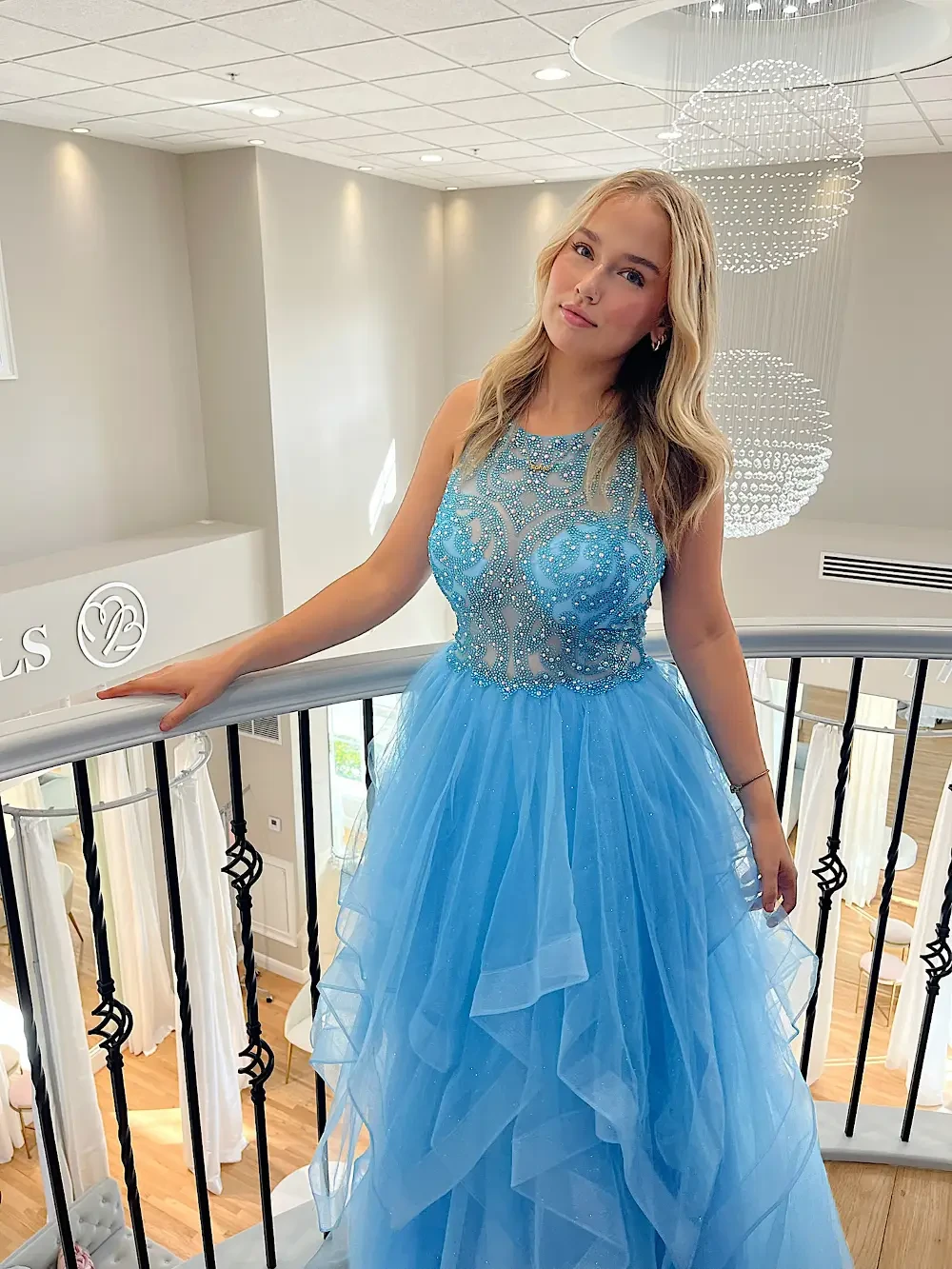 A young woman in a light blue formal dress, standing on a staircase with a decorative chandelier in the background.