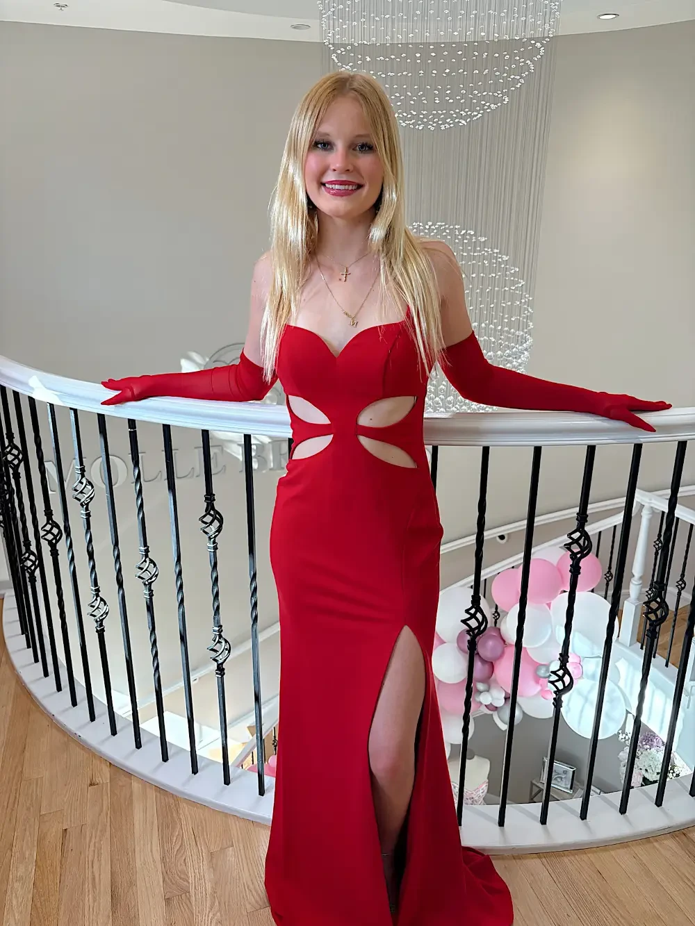 A person wearing a red evening gown with cut-out details and long gloves, standing on a staircase with a chandelier and balloons in the background.