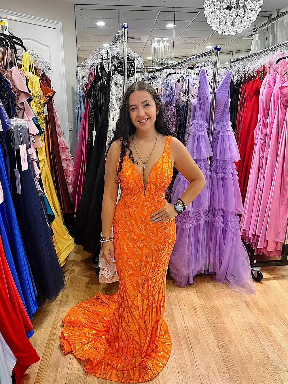 A woman wearing an orange, sequined evening gown stands in a store filled with various dresses.