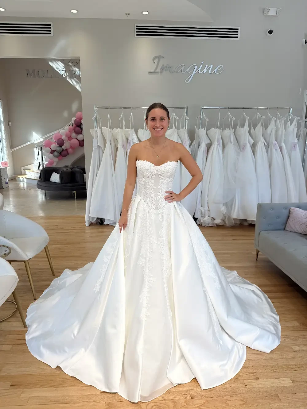 A woman in a white wedding dress stands in a bridal shop, surrounded by hanging white gowns.