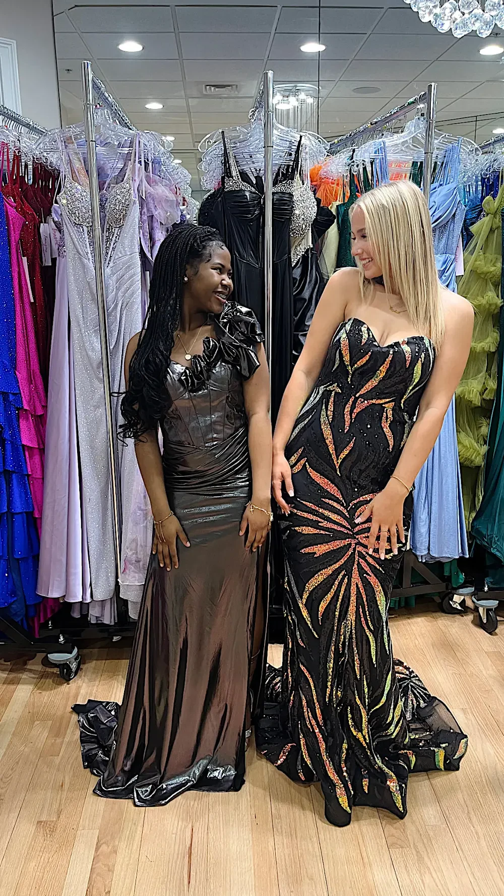 Two women are standing together in a clothing store, surrounded by various formal dresses on racks. One woman is wearing a dark, shiny gown with a floral detail, and the other is in a sequined gown with a colorful, tropical pattern.