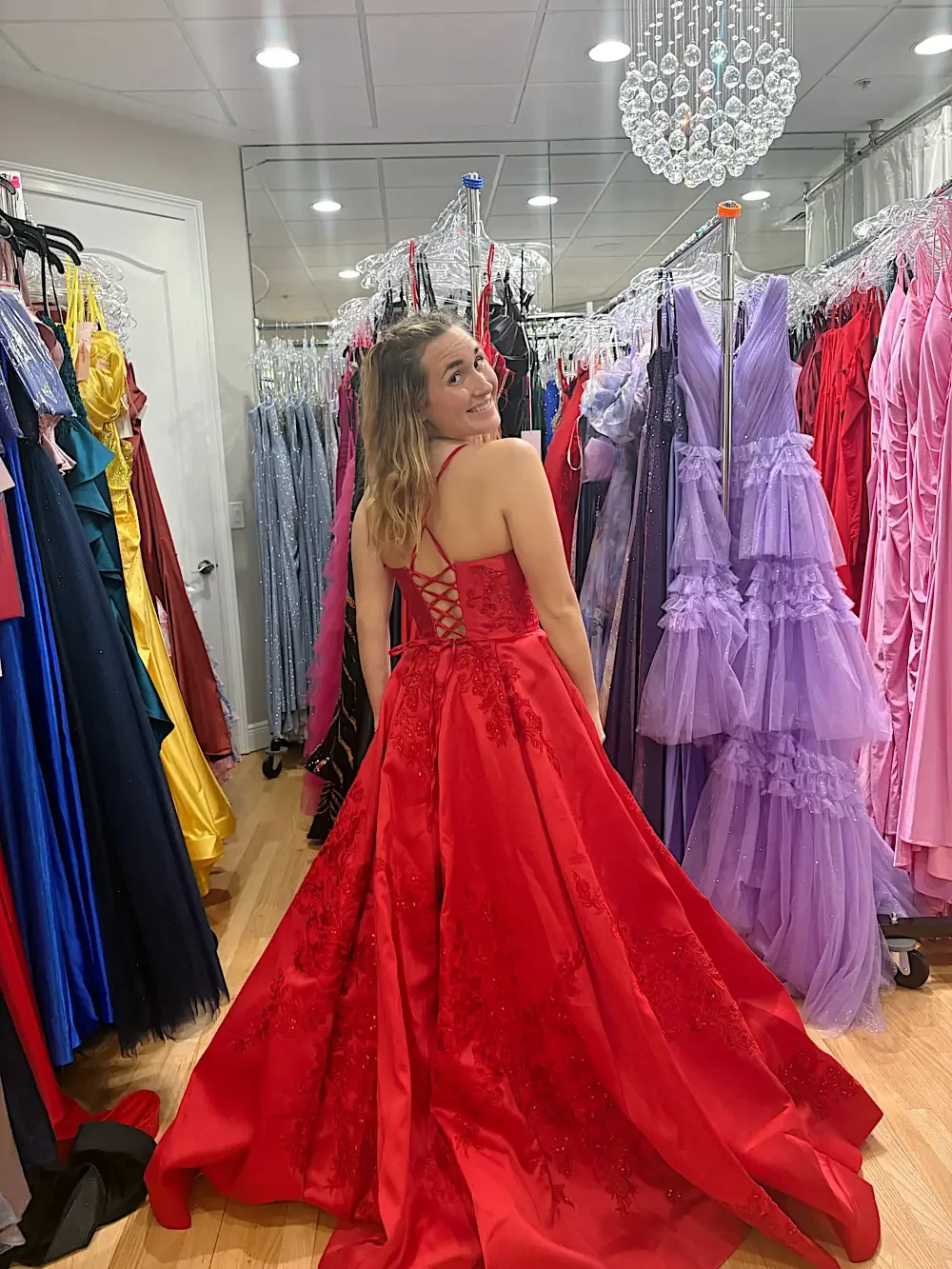 A young woman wearing a red evening gown, standing with her back to the camera in a dress shop surrounded by various colorful dresses.