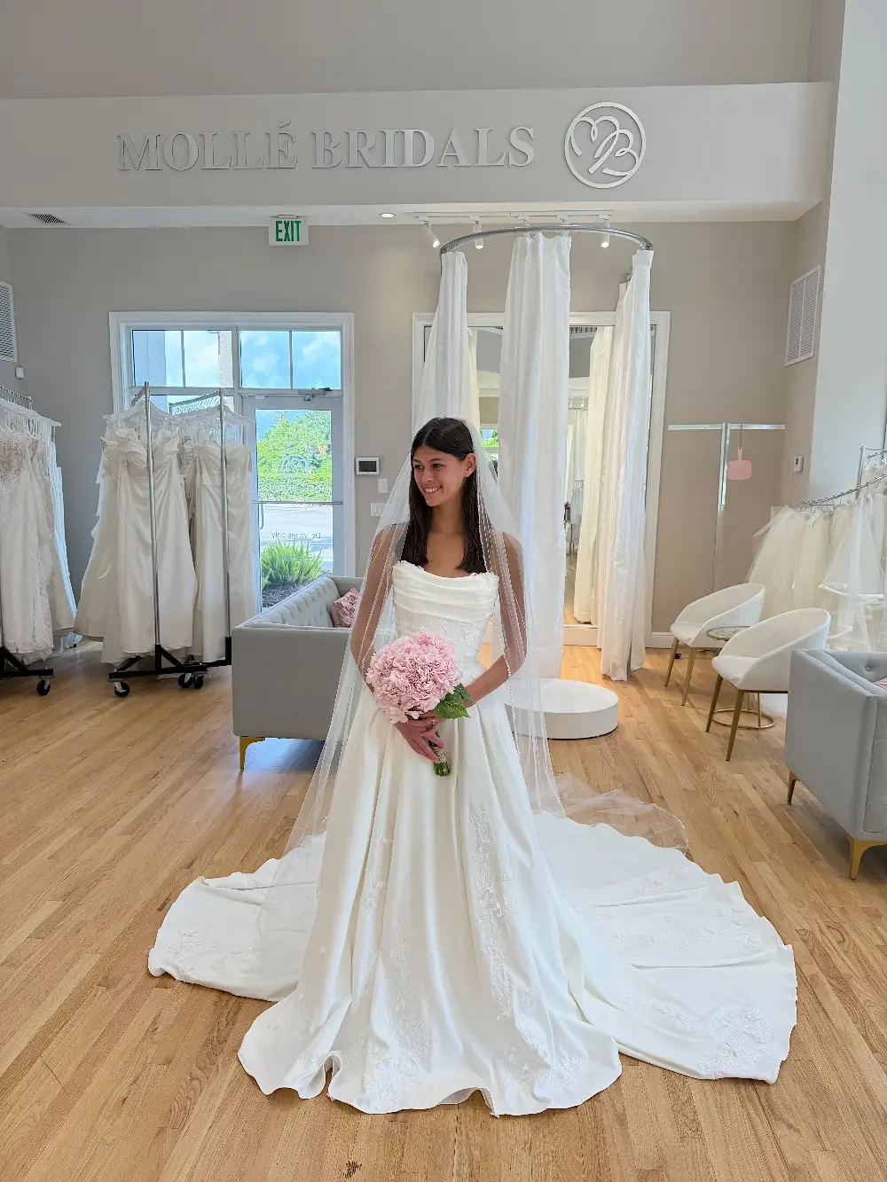 A woman in a wedding dress holding a bouquet of pink flowers in a bridal shop.