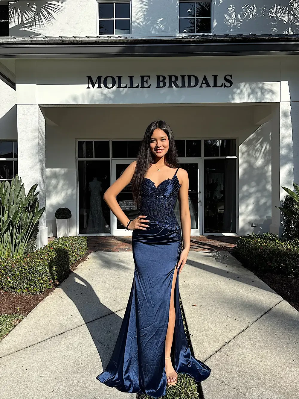 A woman in a form-fitting navy blue gown with a slit stands confidently outside a bridal shop named 