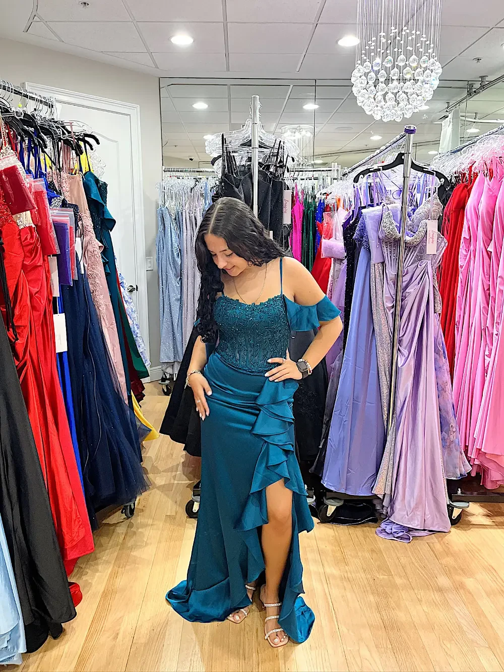 A person trying on a teal dress with ruffles in a boutique filled with various colorful gowns.