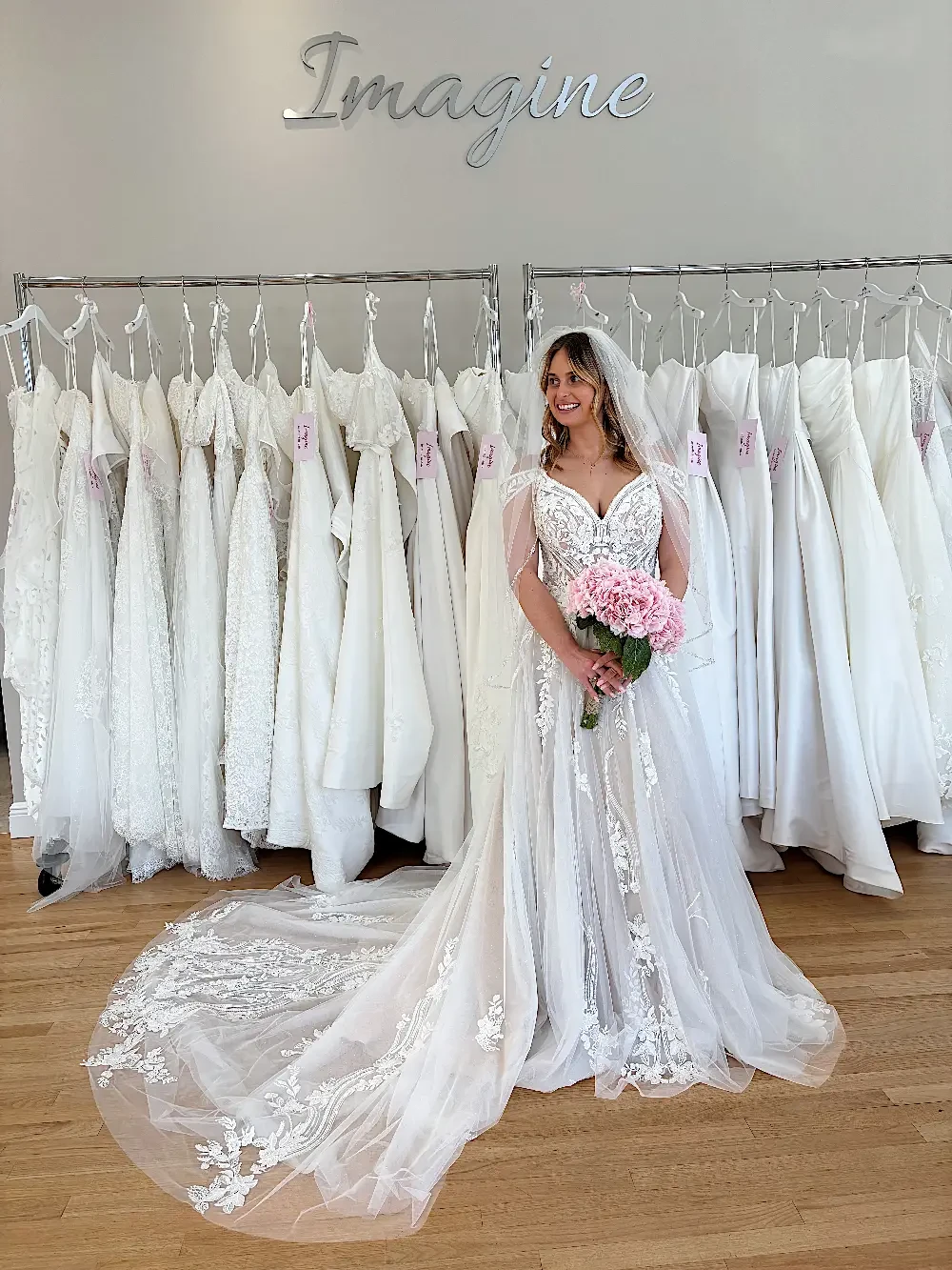 A bride in a wedding dress holding pink flowers stands in front of a rack of white wedding gowns with the word 