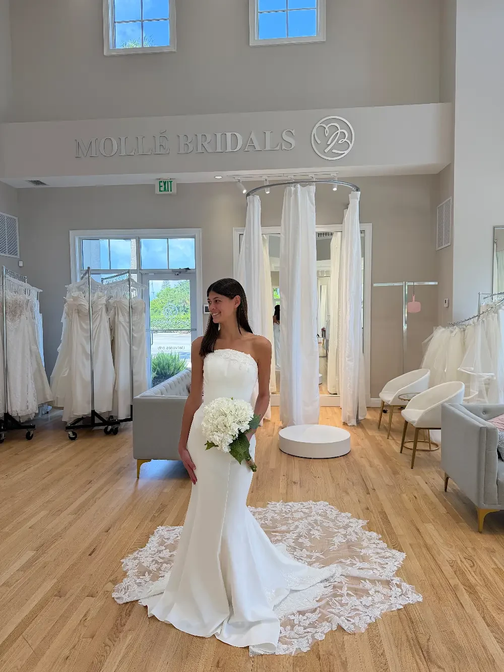 A bride in a white wedding gown poses in a bridal shop, holding a bouquet of white flowers. The shop features wedding dresses displayed on racks and a light, airy interior with large windows.
