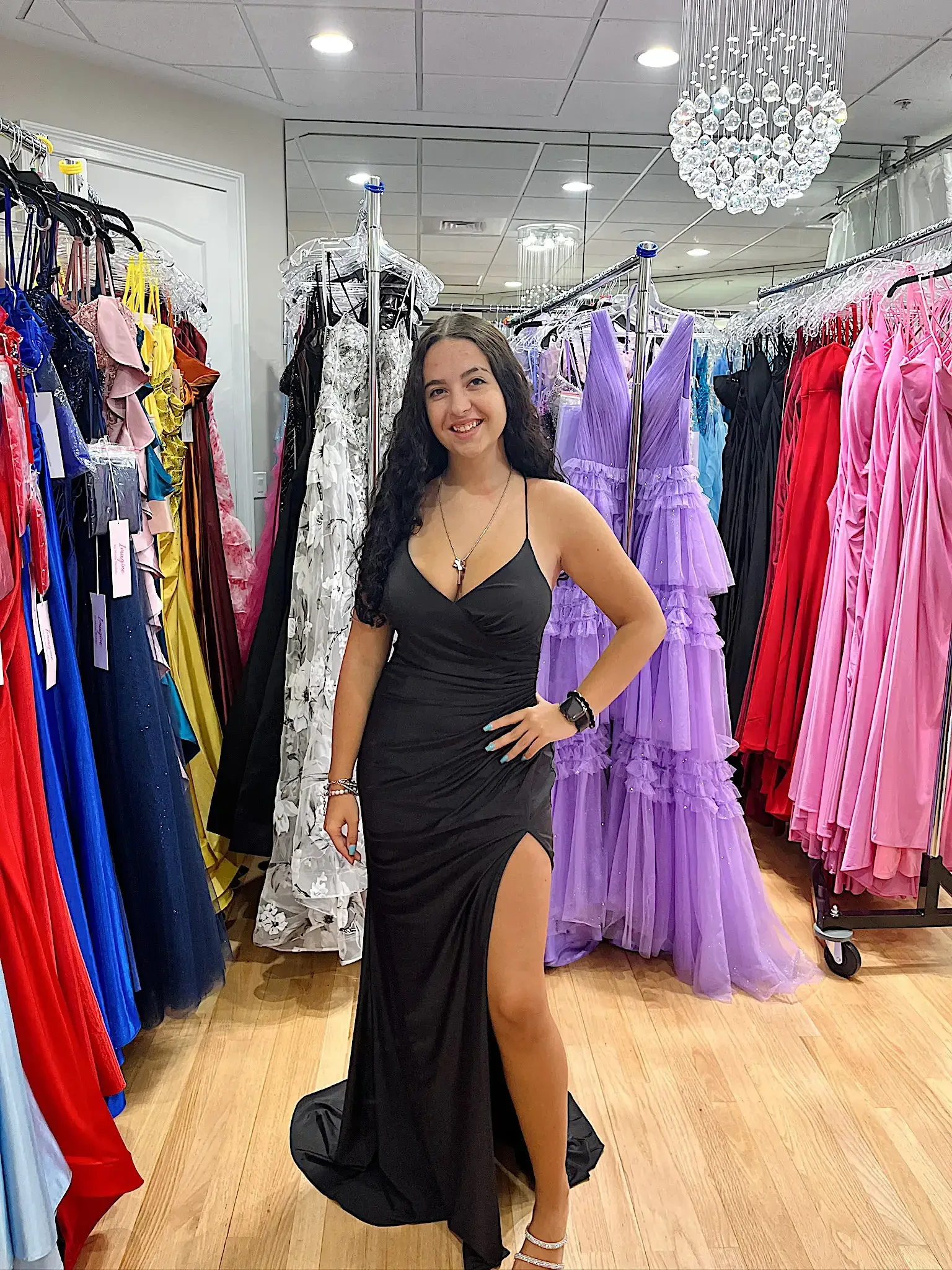 A woman wearing a black dress with a high slit poses in front of a rack of colorful evening gowns.