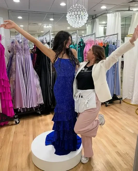 A woman in a sparkling blue dress stands on a pedestal, posing joyfully with another woman who is wearing a blazer and has her arm raised in excitement. They are surrounded by various colorful dresses in a boutique.
